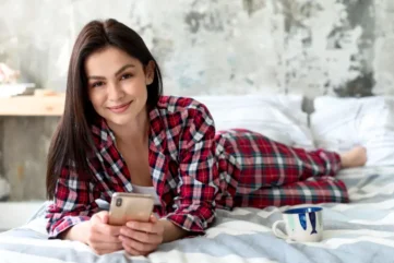 Vrouw op bed in flanellen pyjama met telefoon en kopje koffie.