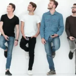 Vier mannen met jeans leunen tegen een witte muur.