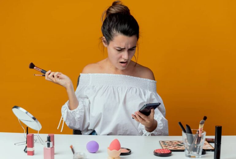 Vrouw kijkt geschrokken op mobiele telefoon met make-up spullen op voorgrond