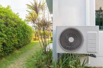 Airconditioner buitenunit naast tuin met groene heggen.