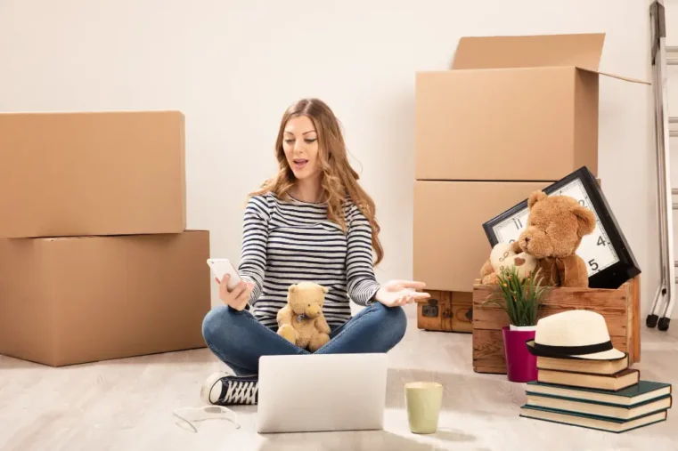 Vrouw met telefoon omringd door dozen en spullen.