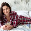 Vrouw op bed in flanellen pyjama met telefoon en kopje koffie.