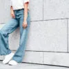 vrouw met jeans en witte sneaker leunt tegen witte muur