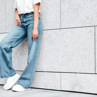 vrouw met jeans en witte sneaker leunt tegen witte muur