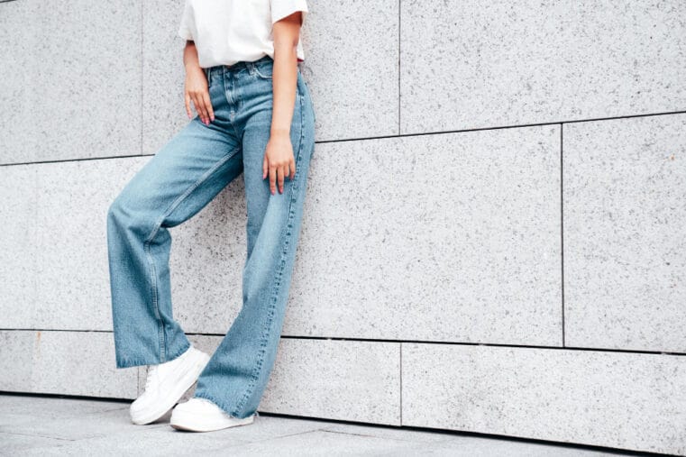 vrouw met jeans en witte sneaker leunt tegen witte muur